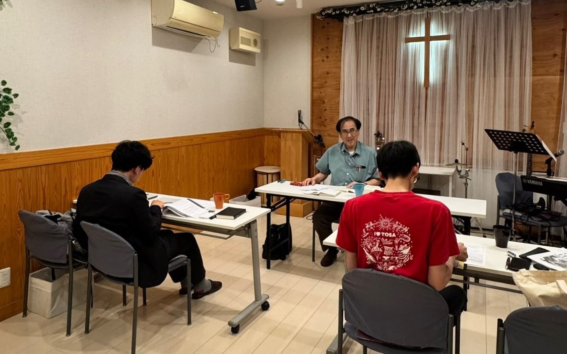 横浜教室で、机を囲んで学ぶ教師と受講者たち。奥の窓には十字架が見える。